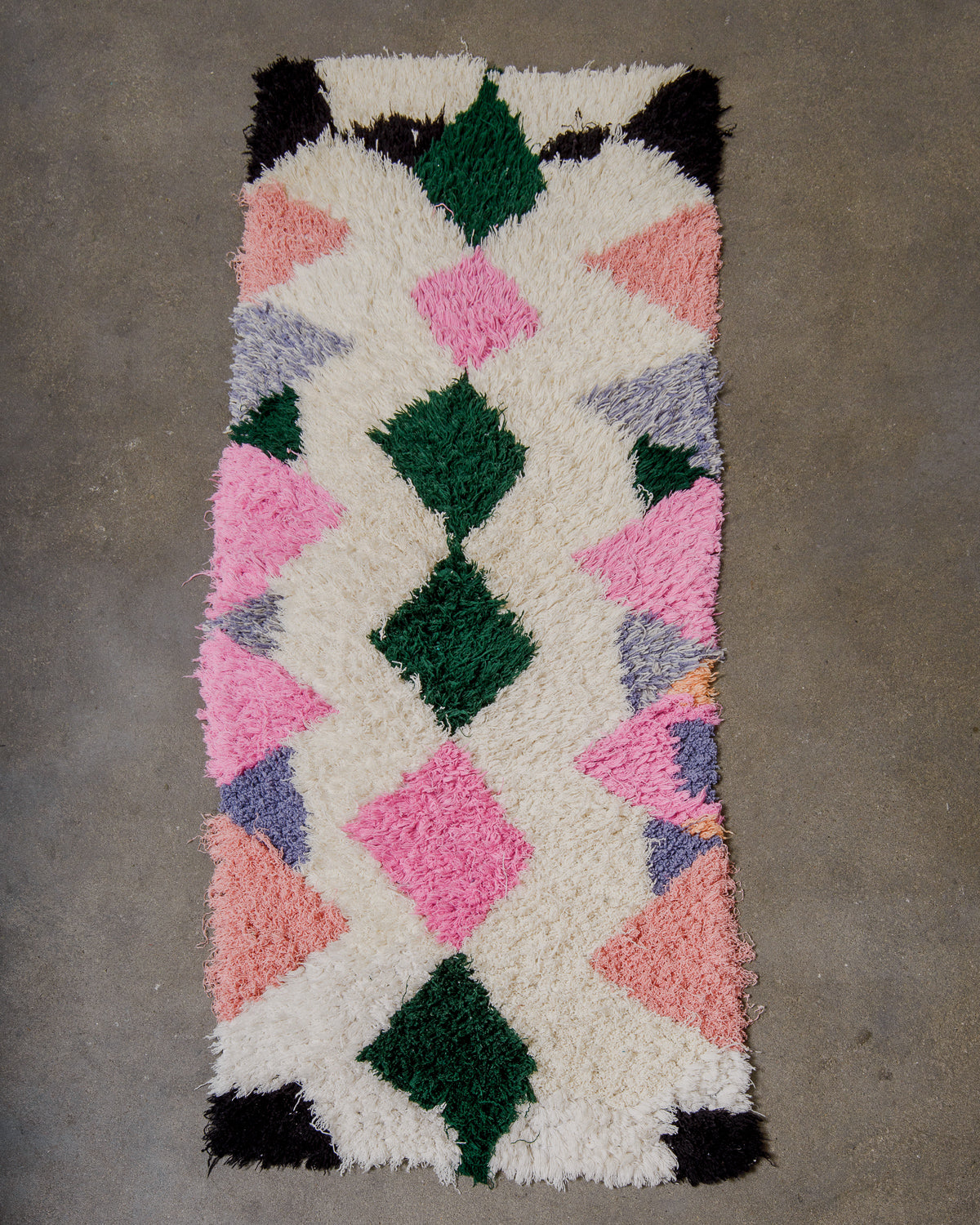 Geometric patterned rug with pink, green, and black diamonds on a cream base, on a cement floor.